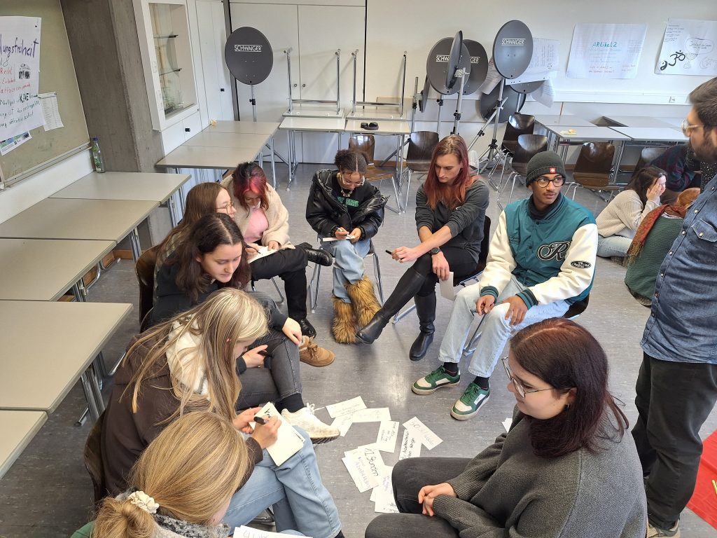 on Schülern und Schülerinnen beschriftete Moderationskarten liegen auf dem Boden; außer herum sitzen SuS im Stuhlkreis.